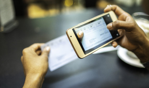 Young woman depositing check by phone in the cafe. Photo courtesy of Getty Images....