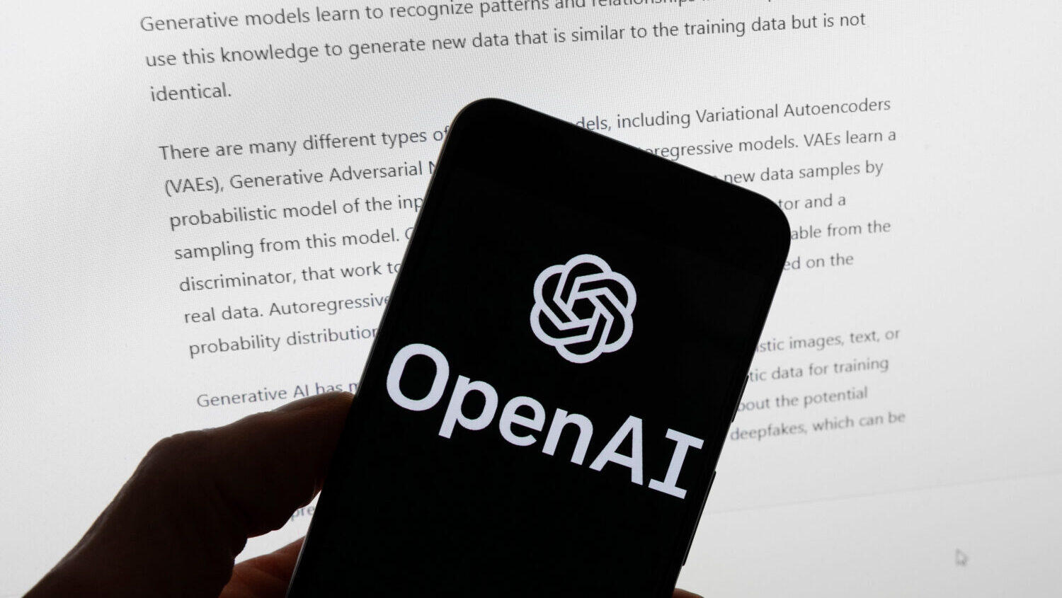 The OpenAI logo is displayed on a mobile phone in front of a computer screen with output from ChatG...