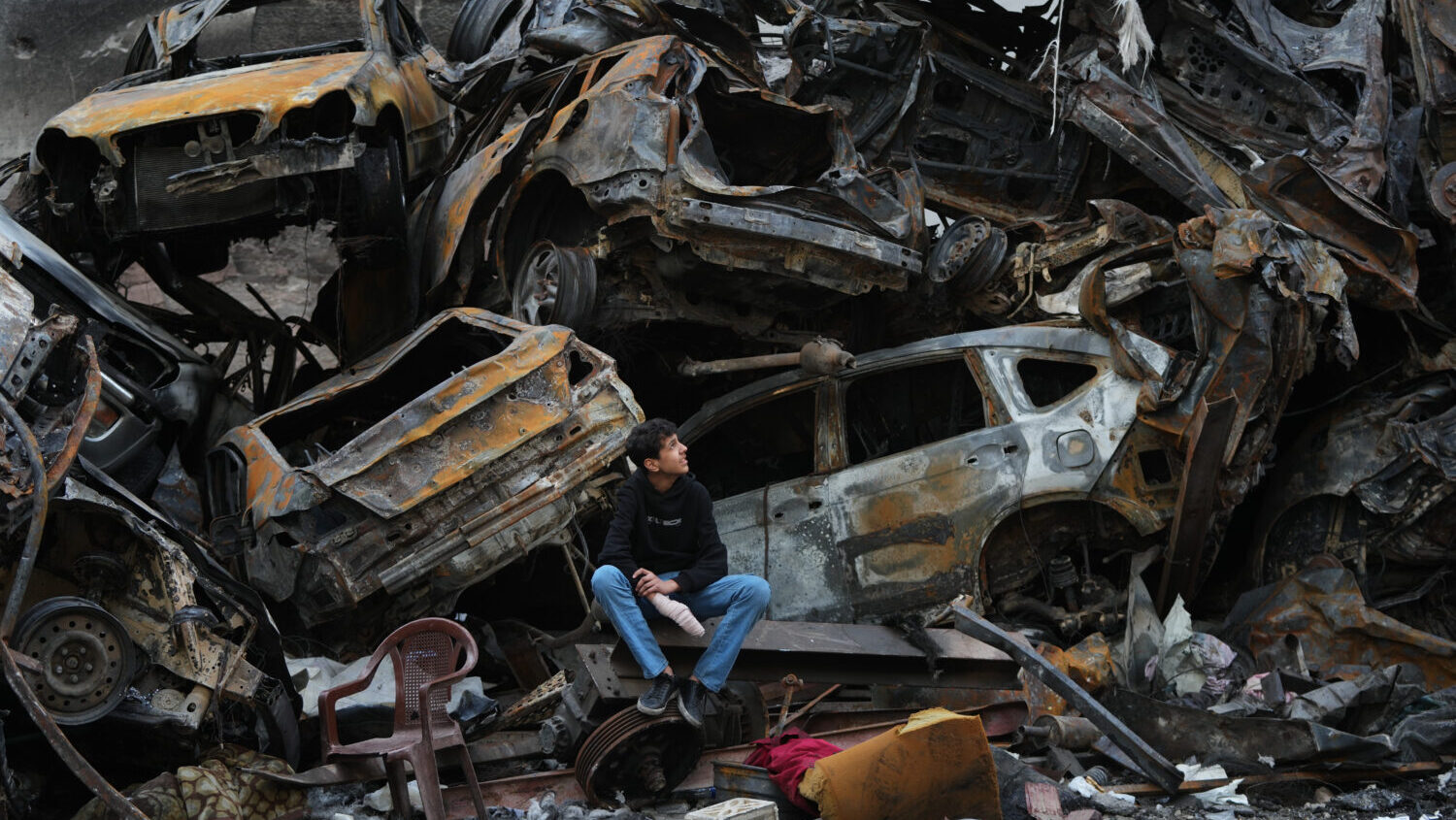 A man sits next to charred cars and wreckage where a building was destroyed by an Israeli airstrike...