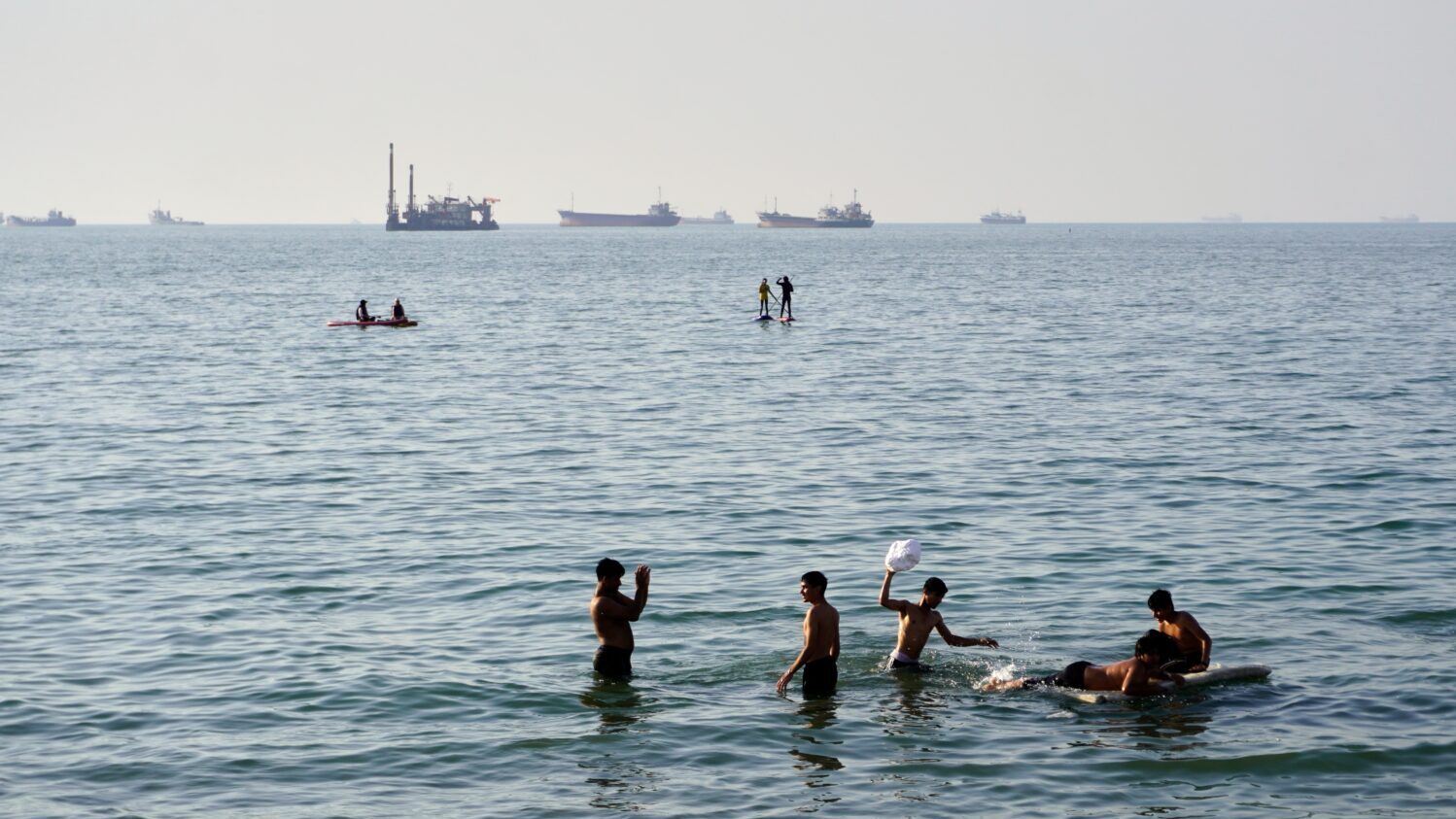 Residents swim and play in the waters of the Strait of Hormuz while cargo ships and commercial vess...